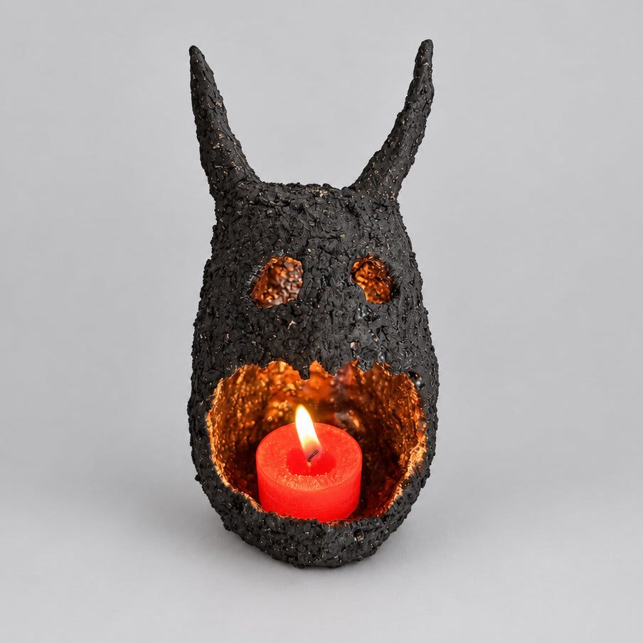 A black textured horned skull candle holder with a glowing red pillar candle inside, set against a plain light beige background.