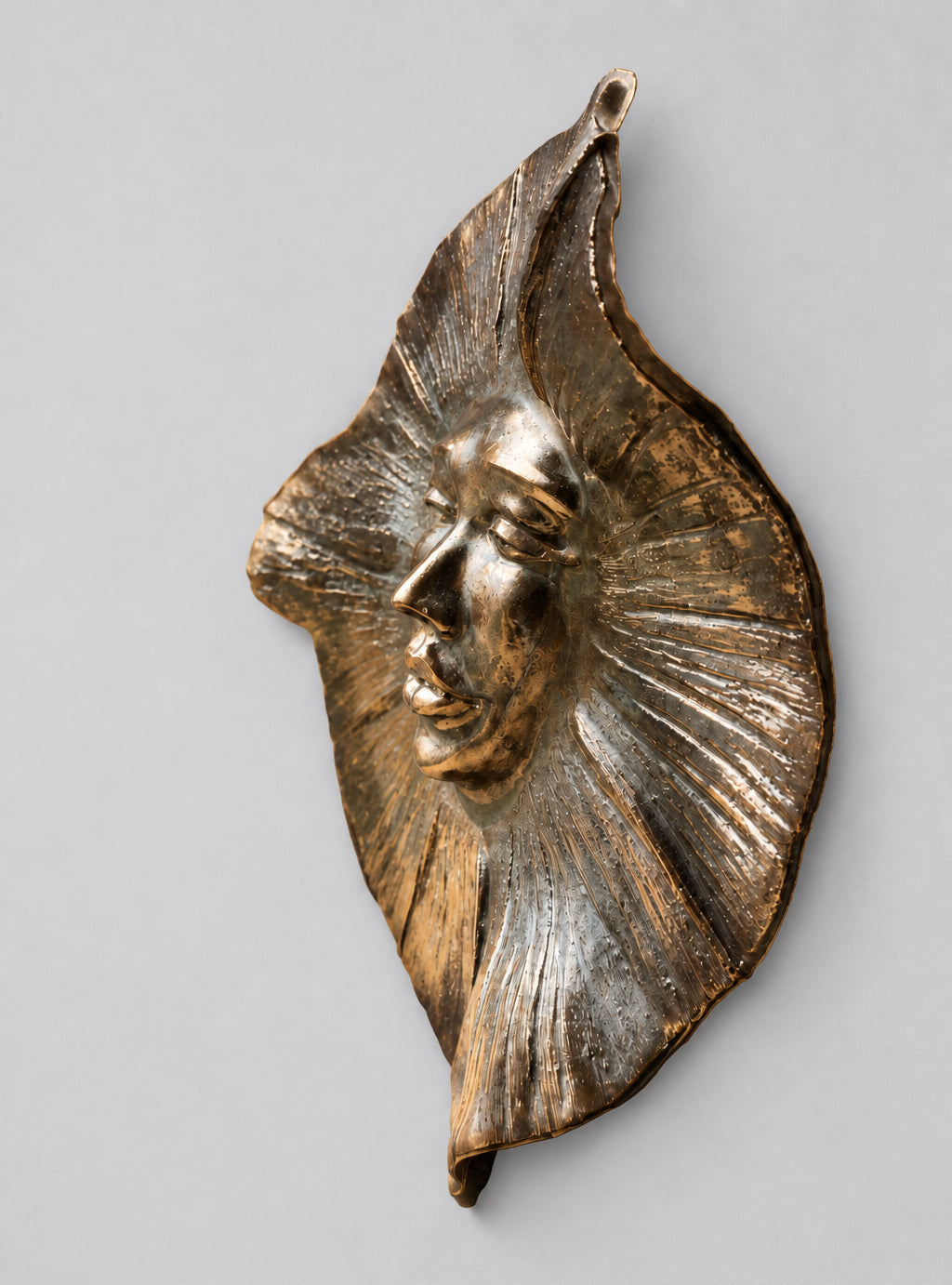 A bronze wall-mounted sculpture of a stylized human face with radiating leaf-like or flame-like extensions, shown against a plain light gray background.