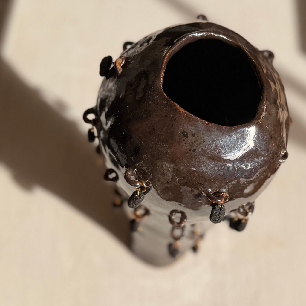 A handmade ceramic vessel with a dark, textured glaze and protruding bead-like details, photographed on a light-colored wooden surface.