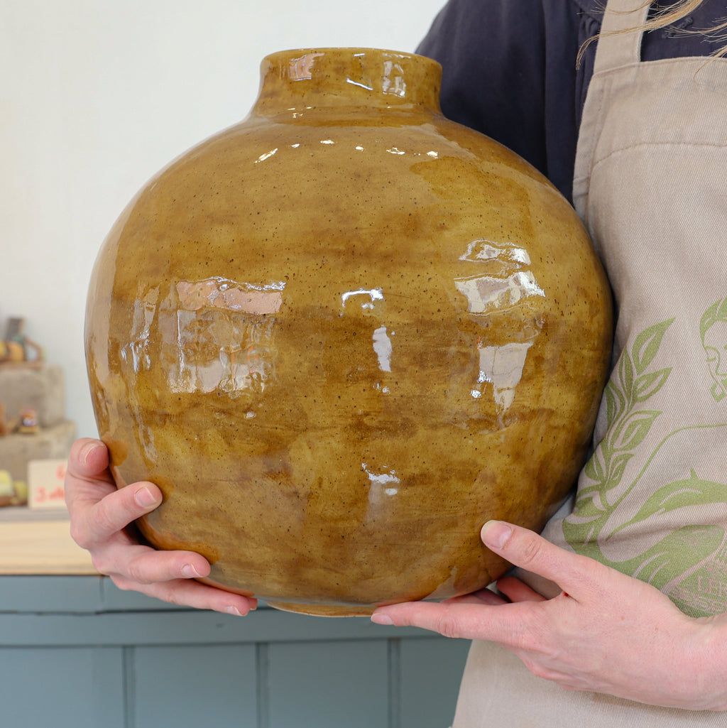 Person holding a large brown ceramic vase with golden green glaze in a workshop setting